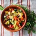 Photo shows aerial view of a bowl of colorful peach salsa on a striped cloth. There is a bunch of cilantro curved around the bowl.