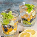 2 glasses filled with oranges, blueberries, mint leaves on a wooden board with orange slices beside the glasses