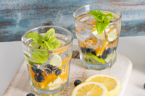 2 glasses filled with oranges, blueberries, mint leaves on a wooden board with orange slices beside the glasses