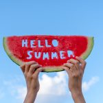 A piece of watermelon against a blue sky. Childrens hands are holding a slice of watermelon with the text Hello Summer. Summer time concept