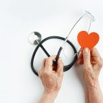 Elderly wrinkled hands holds a stethoscope and heart symbol.