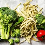 Raw tagliatelle pasta with spinach, broccoli and cherry tomatoes on a grey background.