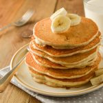 Stack of pancakes with banana slices and honey, on wooden background.