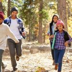 Family of two adults and two children in seasonal clothing run through a forest of autumn foliage