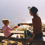 Mother and daughter clinking their mugs at a wooden bench in front of a mountain background