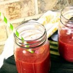 Two mason jars of a bright fuschia drink with bright striped straws sit on a dark table with a brick background