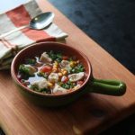 ceramic bowl with long green handle filled with soup on a wooden board with a red and white striped napkin with spoon on top