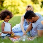 Child and adults laying or sitting on a picnic blanket in the park, eating and laughing