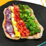 flatbread covered with red onion, yellow squash, red pepper, carrots, green pepper and broccoli on a black plate