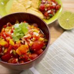 Bowl of red tomato salsa with background of a plate of chips and salsa with lime