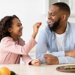 African American father and daughter eating veggies together in the kitchen