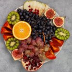 platter of fresh fruits: oranges, grapes, kiwi, persimmons, figs, pomegranate, apple on gray background. view from above