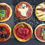 6 small round pieces of dough with tomato sauce on them and decorated with halloween designs made of white cheese, olives and vegetables on a black background