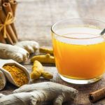 ginger tea in a glass mug with a spoon inside it and 2 scoops of spices with ginger root and a bunch of cinnamon sticks beside it