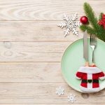 small blue plate with a fork and knife in a santa jacket silverware sleeve with greenery in background on light wooden table