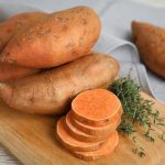 a pile of sweet potatoes next to stack slices of sweet potato and fresh thyme all on a wooden cutting board
