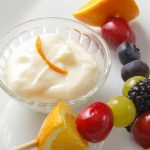 glass bowl with yogurt dip inside and 2 sticks with assorted fruit beside the bowl