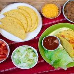 2 Tacos on a plate surrounded by bowls of toppings and a plate of taco shells