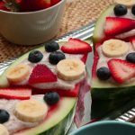 A silver platter with pieces of watermelon covered in yogrut and topped with bananas, blueberries and strawberries
