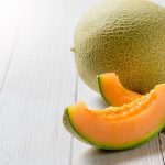 two slices of fresh cantaloupe with the rind intact placed in front of a whole cantaloupe all sitting on a light wooden surface