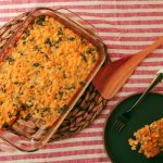 Spicy Kale Corn Pudding in a glass casserole dish with a piece of the corn pudding on a black plate beside the casserole dish