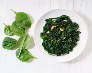 bowl of cooked spinach with garlic, bowl sitting next to uncooked spinach leaves