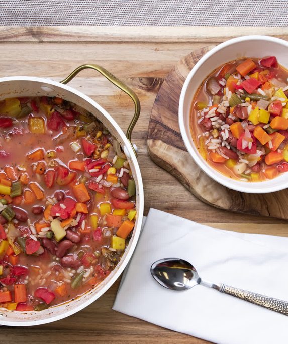 pot of gumbo next to a bowl of gumbo, both sit on top of a cutting board next to a spoon and napkin